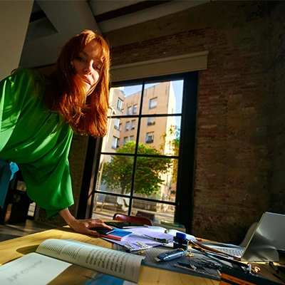 Frau im grünen Hemd steht am Schreibtisch mit Laptop und Büromaterial; Gebäude und Baum durch das Fenster sichtbar.