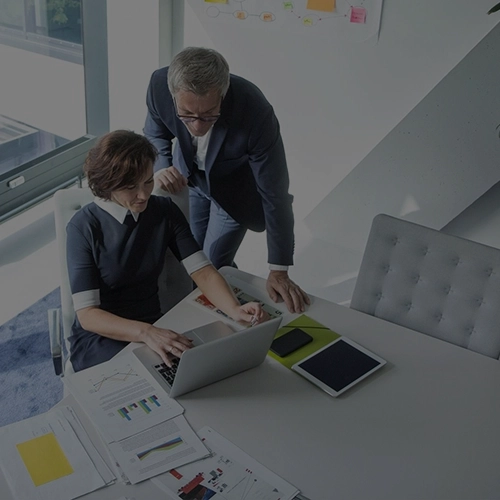 Mann und Frau in Businesskleidung sprechen im Büro vor Laptop; Schreibtisch mit Unterlagen und Stiften.