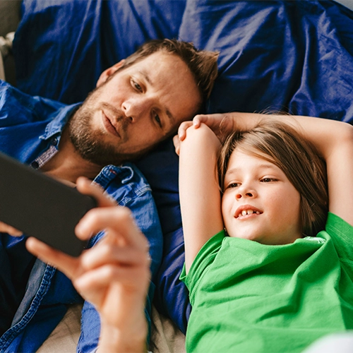 Mann und Kind liegen nebeneinander und schauen gemeinsam auf ein Smartphone.