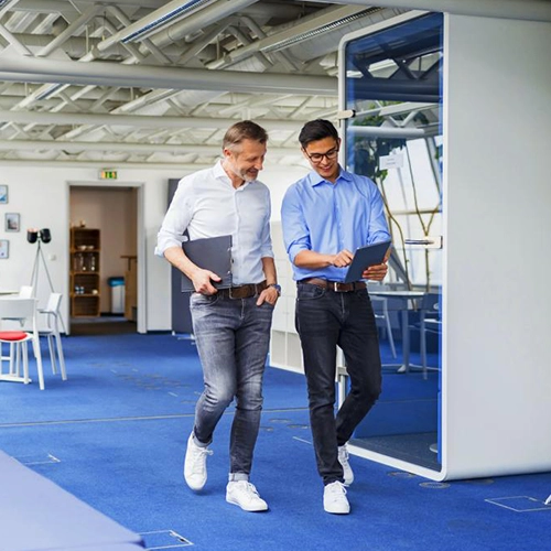 Two men in a modern office and casual attire discussing documents.