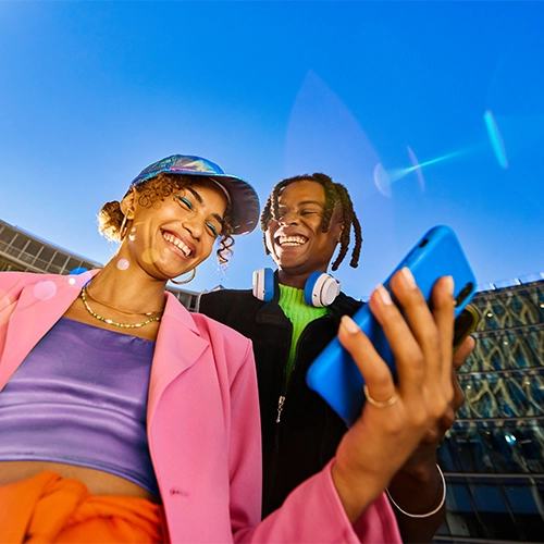 Man and woman in casual clothes hold smartphones and smile; urban environment with blue sky.